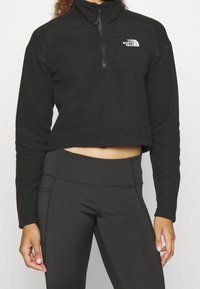 Sudadera de forro polar negra con cremallera, diseño corto, mangas largas y un logo en el lado izquierdo del pecho. Textura suave y costuras mínimas.