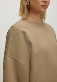 Beige oversized sweatshirt met een ronde hals, geribbelde manchetten en een gladde textuur, gecombineerd met grote, gestructureerde oorbellen.