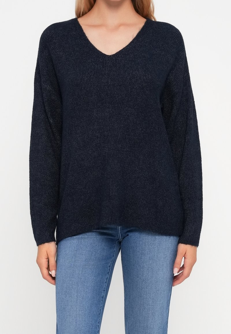 Femme portant un pull bleu marine foncé à manches longues et coupe ample, col en V, avec un jean bleu clair, debout devant un fond uni.