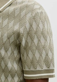 Gestricktes Kurzarmhemd mit beigem und weißem Rautenmuster und geripptem Ärmelbündchen mit weißen Streifen am Arm einer Person.