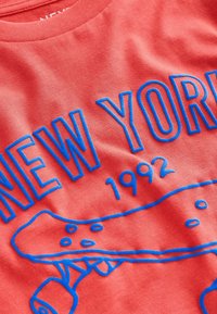Koralli puuvillainen t-paita, jossa on sininen graafinen teksti "NEW YORK 1992" ja skeittauskuvio, pehmeällä pinnalla ja pyöreällä kaula-aukolla.