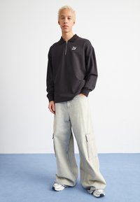 Zwarte sweatshirt met rits en kraag, voorzien van een klein logo op de borst, gecombineerd met losse lichtblauwe cargobroeken en witte sneakers.