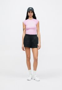 Kobieta stojąca w różowym topie bez rękawów, czarnych szortach, białych tenisówkach, białych skarpetkach oraz czarnej czapce Nike na jednolitym tle.