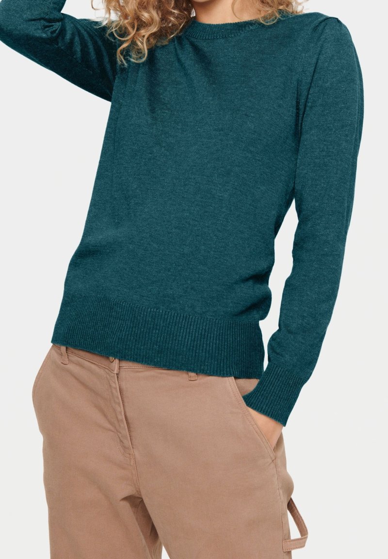 Teal gebreide trui met lange mouwen en ribgebreide manchetten, gecombineerd met lichtbruine broek. De textuur lijkt zacht en comfortabel.