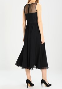 Abito nero midi con corpino aderente, sovrapposizione semi-trasparente a pois sul retro e gonna fluida. Indossato con scarpe con tacco alto nere.