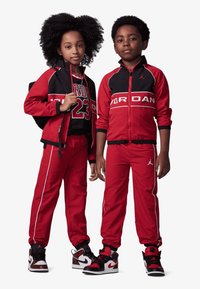 Rote und schwarze Sportanzüge mit weißen Akzenten. Kinder tragen passende Jacken mit Reißverschluss vorne und Logo, kombiniert mit sportlichen Schuhen und Rucksack.
