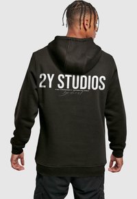 Felpa nera con cappuccio, caratterizzata da grande testo bianco "2Y STUDIOS" sulla schiena, insieme a ulteriore testo più piccolo sottostante.