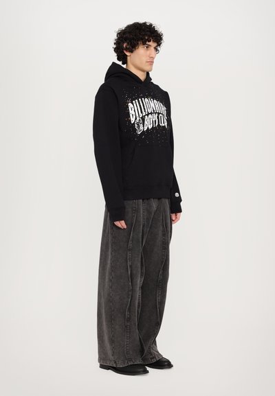 Hoodie noir avec un graphique sur le devant "Billionaire Boys Club" et des accents colorés, associé à un pantalon en denim gris ample et à des chaussures noires.