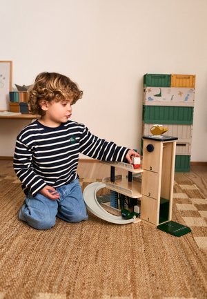 Enfant aux cheveux bouclés portant un t-shirt rayé, jouant avec un garage jouet en bois à plusieurs niveaux et des véhicules sur un sol moquetté à l'intérieur.