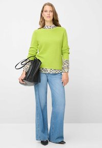 Maglione verde a coste indossato sopra una camicia a fantasia, abbinato a jeans svasati azzurri e una borsa a secchiello nera. Texture lisce, stile casual.