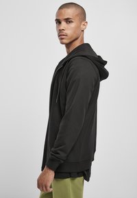 Sudadera con cremallera negra, capucha con cordón, textura suave, puños acanalados y silueta ajustada. Lleva pantalones verde oliva.