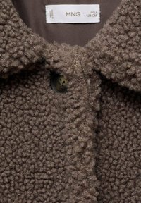 Smeđi flis kaput s teksturiranom površinom, zaobljenim ovratnikom i kopčanjem na dugme. Oznaka sadrži informacije o veličini i održavanju.