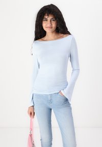Lyseblå langermet topp med off-shoulder-design, laget av et glatt materiale. Bært med lyseblå jeans og en rosa veske.