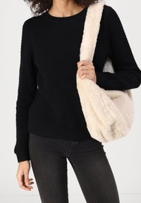 Jersey de punto negro texturizado con puños acanalados, combinado con un bolso de hombro beige y esponjoso. La modelo lleva vaqueros oscuros y sostiene el bolso con una mano.