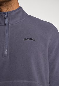 Κοντινή λήψη ενός ατόμου που φοράει ένα βιολετί φλις πόλο με φερμουάρ ενός τετάρτου και το μαύρο λογότυπο "BORG" κεντημένο στο στήθος.