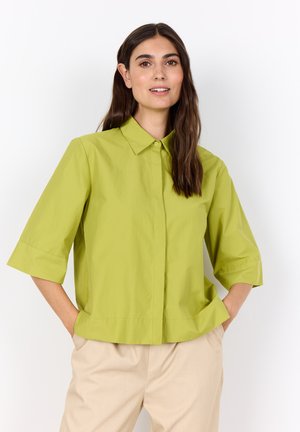 Femme aux longs cheveux bruns portant une chemise verte clair à boutons avec des manches jusqu'au coude et un pantalon beige, les mains dans les poches.