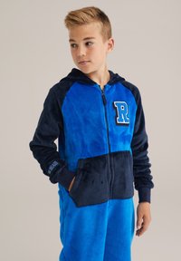Blå fleeceoverall med huva, framdragkedja och kontrasterande marinblå ärmar. Inkluderar en "R"-logotyp och sidofickor. Mjuk textur.