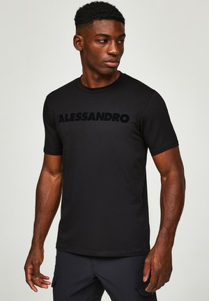Mann trägt ein schwarzes Kurzarmhemd mit der Aufschrift "ALESSANDRO" auf der Brust und schaut nach links vor einem schlichten Hintergrund.