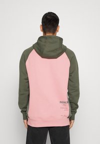Man med kort mörkt hår som bär en rosa och olivgrön Nike-hoodie, vänd bort från kameran mot en enkel ljusgrå bakgrund.