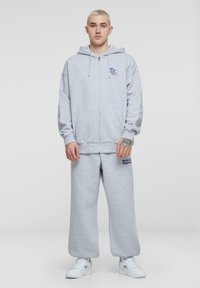 Grå zip-up hoodie og matchende sweatpants i et blødt stof. Hoodie har logo på brystet; bukserne har broderet tekst på siden.
