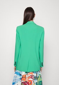 Blazer verde con vestibilità sartoriale, caratterizzato da un colletto a risvolto, maniche lunghe e dettagli con bottoni neri. Tessuto testurizzato e spacco centrale sul retro.