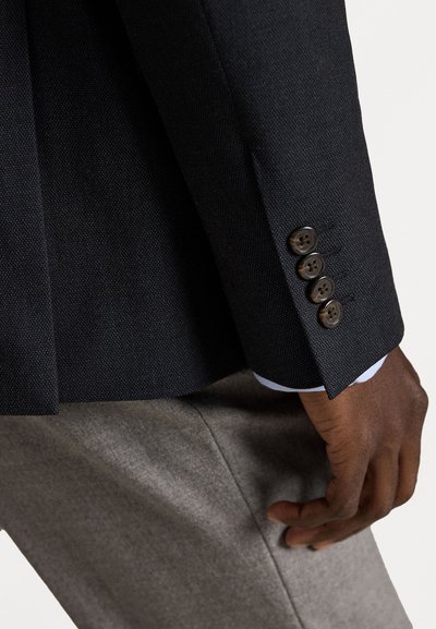Σκούρο υφασμάτινο μανίκι σακακιού με τέσσερα καφέ κουμπιά, σε συνδυασμό με γκρι παντελόνι. Ορατό μανίκι και μπλε πουκάμισο από κάτω.