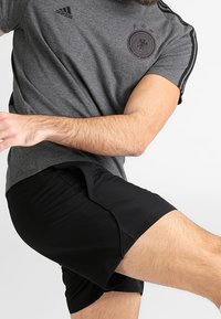 Harmaa urheilupaita mustilla raidoilla, jossa on pyöreä logo, sekä mustat shortsit. Kankaan pinta näyttää sileältä ja rentolta.