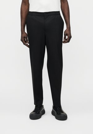 Άνδρας με στενά μαύρα παντελόνια φόρμας, λευκό πουκάμισο σκουπισμένο μέσα, και μαύρα αθλητικά χαμηλού προφίλ που στέκεται μπροστά σε απλό φόντο.