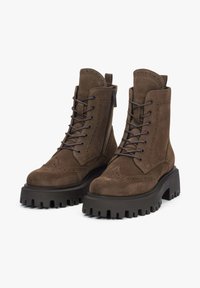 Brown suede ankle boots featuring a chunky rubber sole, lace-up front, decorative brogue detailing, and side zipper.