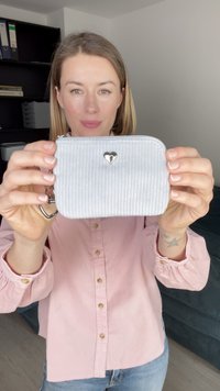 Lichtgrijze corduroy portemonnee met een zilveren hartaccent, ritssluiting en een sleutelhangerbevestiging. Gladde textuur met verticale ribbing.