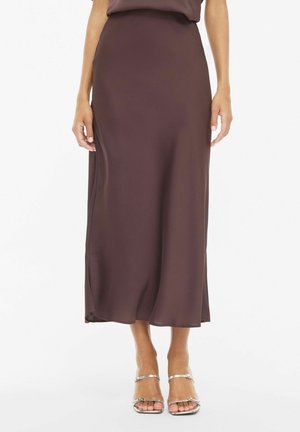 Modèle portant une jupe midi marron foncé taille haute et des sandales argentées à brides, sur un fond de studio blanc.