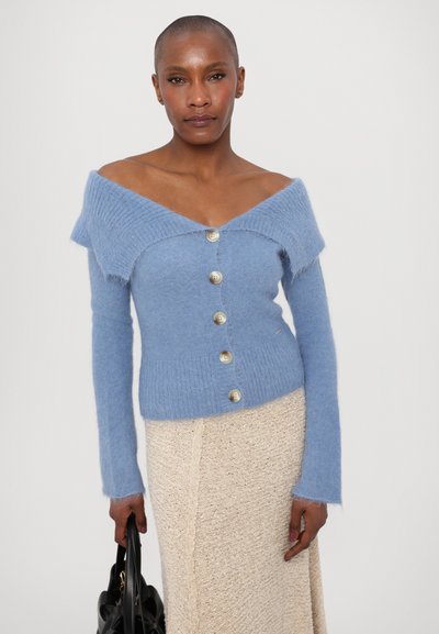 Light blue off-shoulder cardigan with a textured finish, featuring large buttons and long sleeves. Paired with a cream knit skirt.