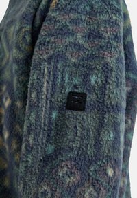 Fleecejacke in Blau- und Grüntönen, mit einem strukturierten Stoff mit einem kreisförmigen Muster und einem schwarzen Markenlogo-Patch auf dem Ärmel.