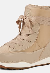 Beżowy botek za kostkę wykonany z pikowanego materiału, z zaokrąglonym noskiem, szerokimi sznurowadłami oraz teksturowanymi akcentami z boku i na podeszwie.