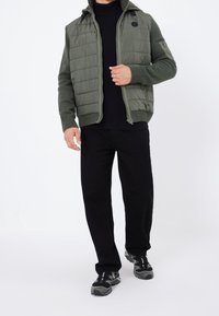 Gilet matelassé vert olive avec fermeture éclair à l'avant et capuche, associé à un pull en maille noir et un pantalon noir, porté avec des chaussures de sport noires.