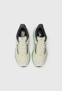 Zapatillas de running beige con upper de malla texturada, acentos verdes, suelas verde claro y forro interior negro. Cordones y detalles del logo visibles.