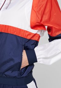 Ręka w kieszeni sportowej kurtki przeciwwiatrowej w kolorze białym, czerwonym i granatowym, z kieszenią na zamek.