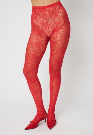Jambes portant des collants en dentelle florale rouge associés à des talons aiguilles rouges à bout pointu sur un fond uni.