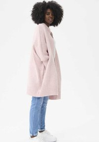 Różowy, oversize'owy sweter z dzianiny z prążkowanym dołem; noszony na białej bluzce, zestawiony z jasnoniebieskimi dżinsami i białymi trampkami.