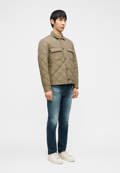 Άνδρας στέκεται φορώντας μια καπιτονέ χακί ζακέτα με κουμπιά, μπλε τζιν και λευκά αθλητικά παπούτσια μπροστά σε απλό λευκό φόντο.