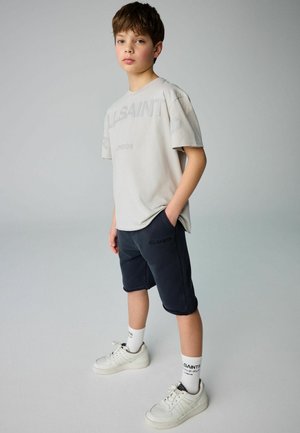 Ljusgrå bomullströja med präglad text "ALLSAINTS LONDON", i kombination med marinblå shorts och vita sneakers med svarta accenter samt logosockor.