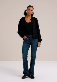 Črna corduroy jakna z ribasto teksturo, v kombinaciji s črnim topom in modrimi džins hlačami. Model nosi črne superge in bele okrogle uhane.