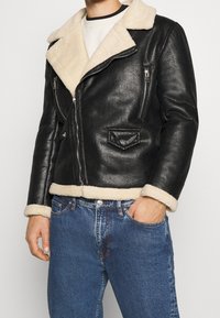 Czarna skórzana kurtka z kremowym kołnierzem i mankietami z owczej wełny, wyposażona w kieszenie na zamek i zamek błyskawiczny z przodu. Klasyczny design z teksturowanym wykończeniem.