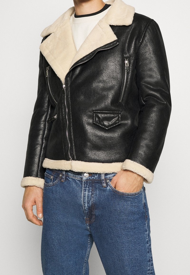 Czarna skórzana kurtka z kremowym kołnierzem i mankietami z owczej wełny, wyposażona w kieszenie na zamek i zamek błyskawiczny z przodu. Klasyczny design z teksturowanym wykończeniem.