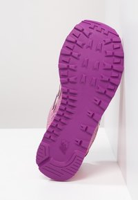 Lila gummisul av en rosa träningssko med djupa mönster, placerad uppstående mot en vit hörnbakgrund.