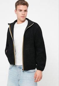 Giovane uomo che indossa una giacca in pile nera con zip e dettagli a righe sopra una maglietta bianca e jeans azzurri chiari, in piedi contro uno sfondo bianco.