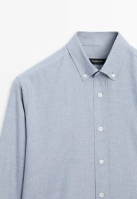 Chemise bleu clair à manches longues avec col, boutons blancs et une étiquette de marque noire visible à l'intérieur du col.