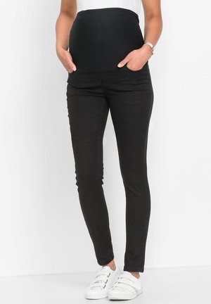 Jeans da maternità neri con vita elastica, design slim fit e tasche. Abbinati a sneaker bianche slip-on.