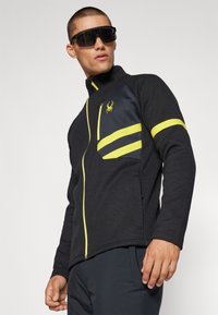 Veste zippée noire en tissu texturé, avec des accents jaunes sur les poches et les manches, arborant un logo d'araignée sur la poitrine.