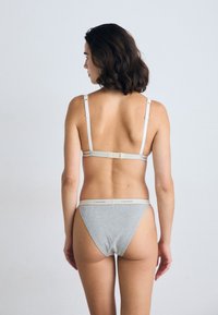 Vrouw met schouderlang donker haar, gekleed in grijze Calvin Klein ondergoed, staande met haar gezicht naar een effen lichtgrijze achtergrond.